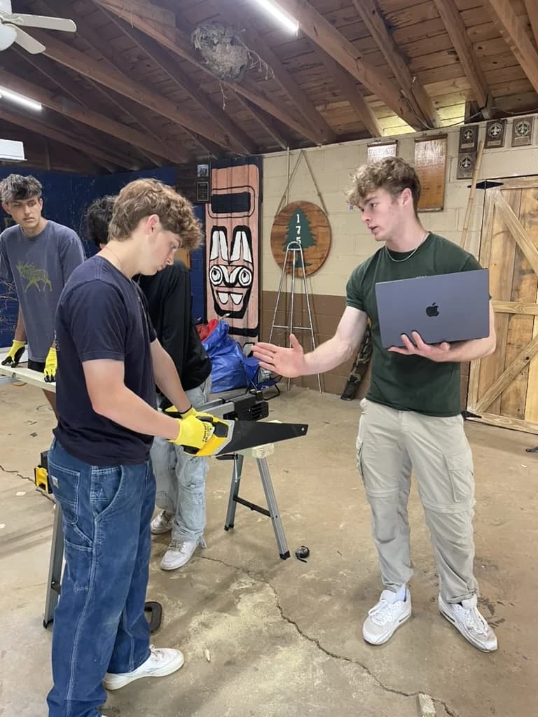 Ethan, holding a laptop, walks a younger scout through a cut at the sawhorses during his Eagle project.
