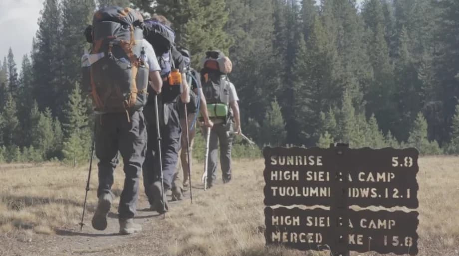A line of scouts with full backpacks and trekking poles hiking past a High Sierra Camp trail sign in Yosemite.